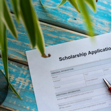 Showing paper application for scholarship on blue, wooden table with plant and with red pen on top of application form.
