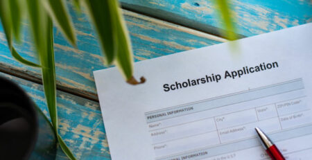 Showing paper application for scholarship on blue, wooden table with plant and with red pen on top of application form.
